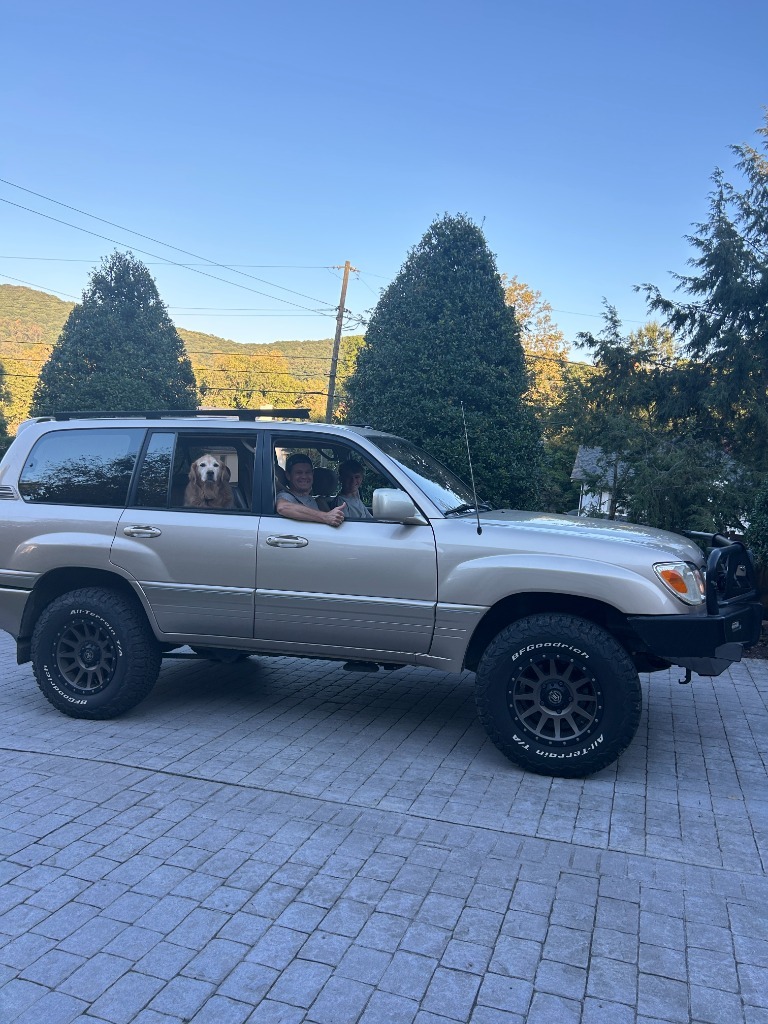A smiling client giving a thumbs up from the driver seat of a clean champagne 100 Series Land Cruiser, with his son in the passenger seat and golden retriever in the back